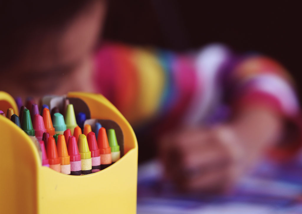 Box filled with colorful crayons, with a child drawing in the background