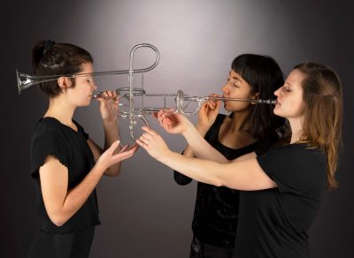 three women blowing a shared glass trumpet